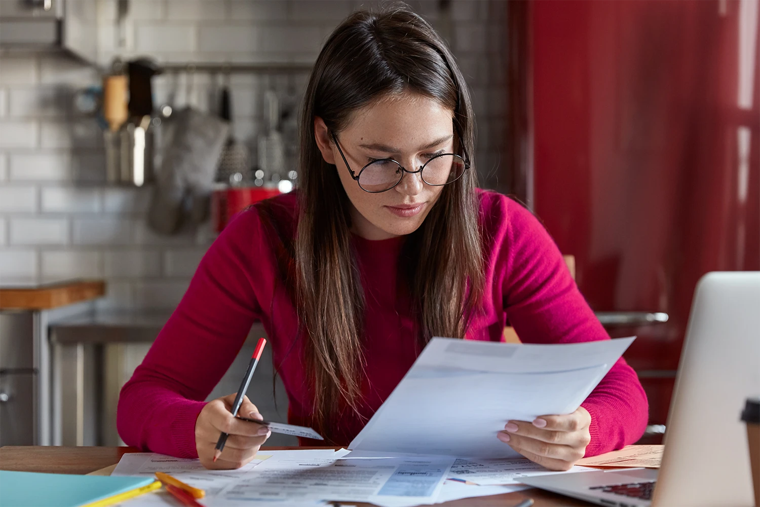 Mulher de blusa vermelha, analisa documentos sentada à mesa, de frente para um laptop e com um lápis em sua mão. A cena exemplifica a importância da regularização de empresas, que consiste no cumprimento de todas as obrigações legais e fiscais.